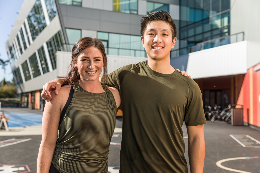 Two students wearing athletic clothing stand outdoors with their arms around each other's shoulders.