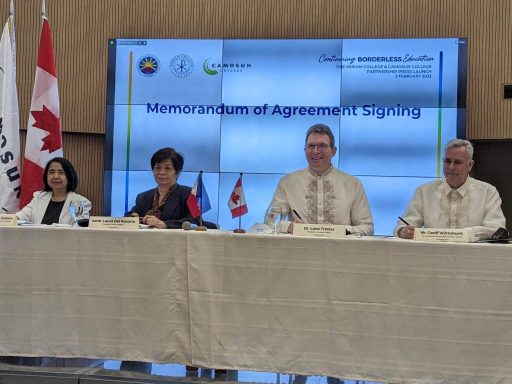 Four people holding pens in hands sit behind a table with a white cloth draped over it and smile for the camera. A digital sign behind them states: "Memorandum of Agreement Signing"