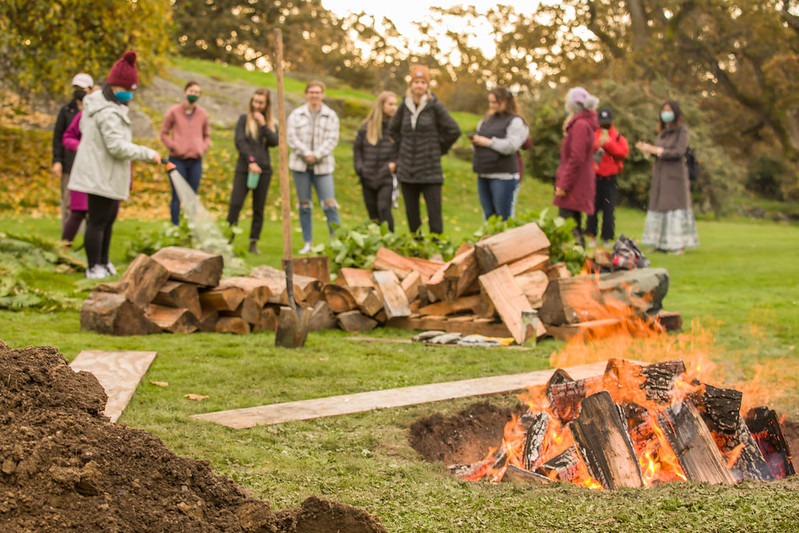 A crowd of people standing outdoors on a fall day with a fire in the ground in the foreground