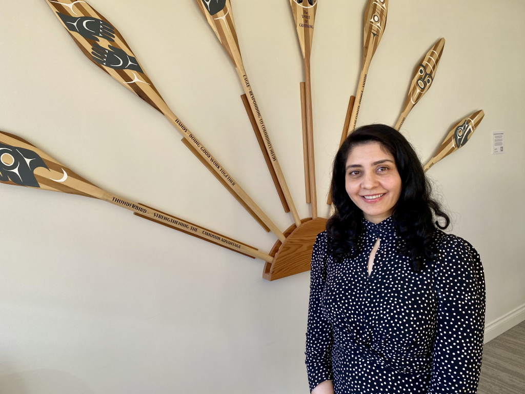 A woman with dark hair stands in front of six carved paddles with Indigenous designs.