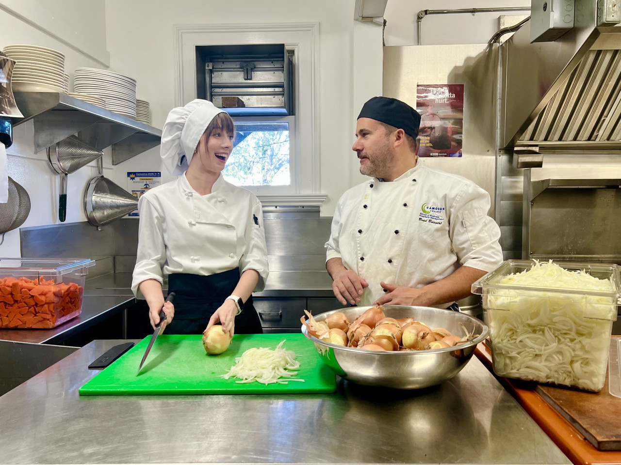 A young women in kitchen whites stands in front of a chopped onion on a green cutting board while talking with an older male in whites. There is a large bowl of onions next to a white container of chopped onions in the foreground.