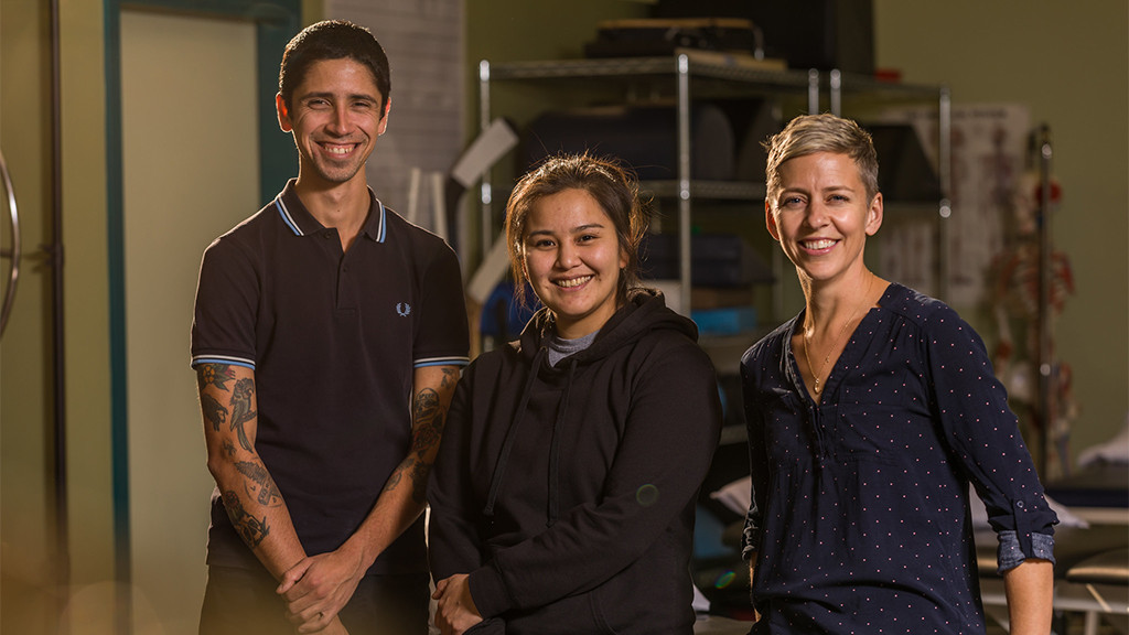 two massage therapy students standing and smiling with instructor in a classroom lab at the interurban campus