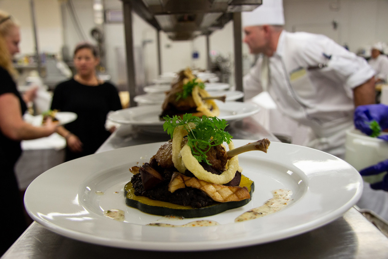A beautifully prested plate with a lambchop sits on the warming counter in a professional kitchen. Chefs are working in the background. 