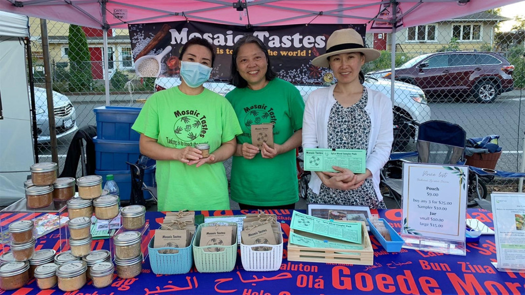 Three women in green shirts reading "Mosaic Tastes" stand at a table with a display of spice blends.