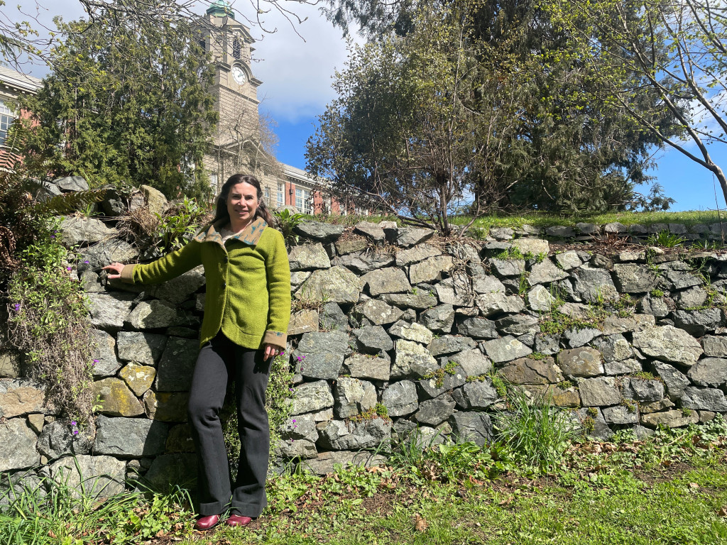 Instructor Nicole Kilburn stands against a stone wall on campus in spring sunlight