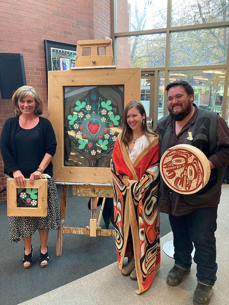 Three people - a woman, a young woman and a man - stand next to a painting on a easel. The painting has what appears to a strawberry, green lights against a dark Indigenous design.