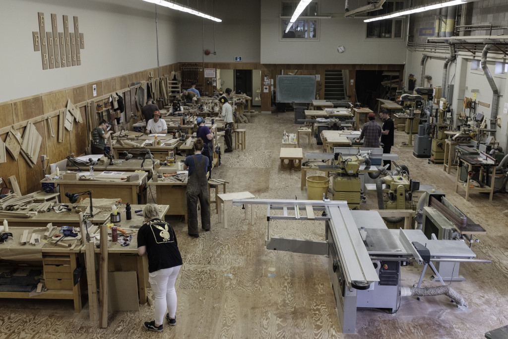 A class scene of students in a large wood shop, head down, working at their stations.
