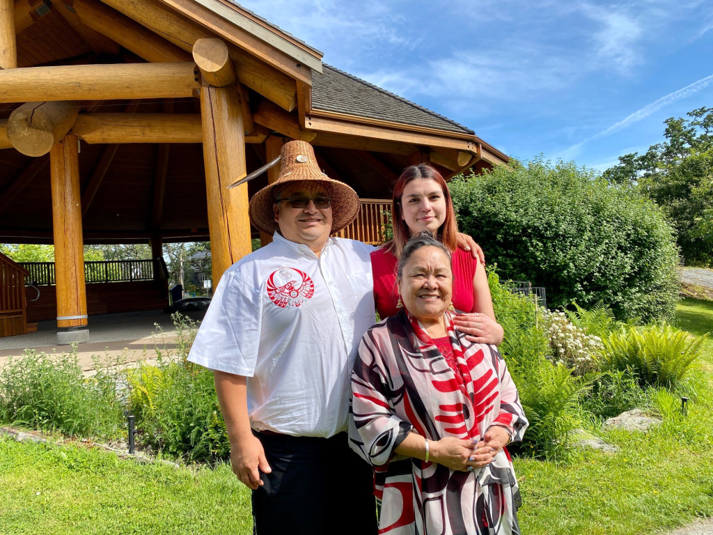 A man and two women stand in front of a traditional Indigenous building