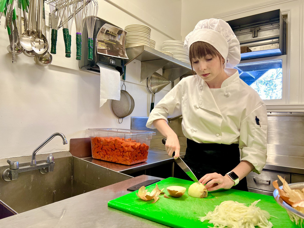 A student wearing white kitchen uniform chops an onion on a green cutting board.