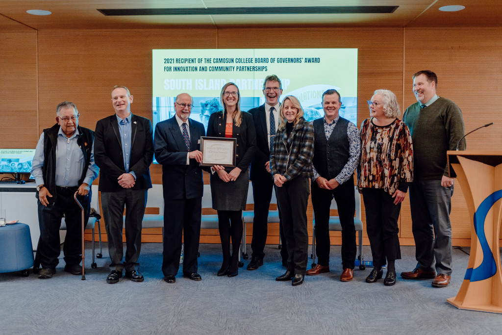 An indoor mixed group of nine people standing in front of a wood panelled wall hold up a framed award