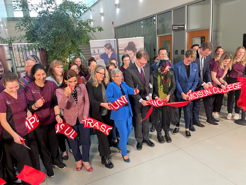 Students wearing scrubs are joined by faculty and BC Cabinet Ministers to cut a ribbon indoors marking the opening of the new campus ultrasound clinic.