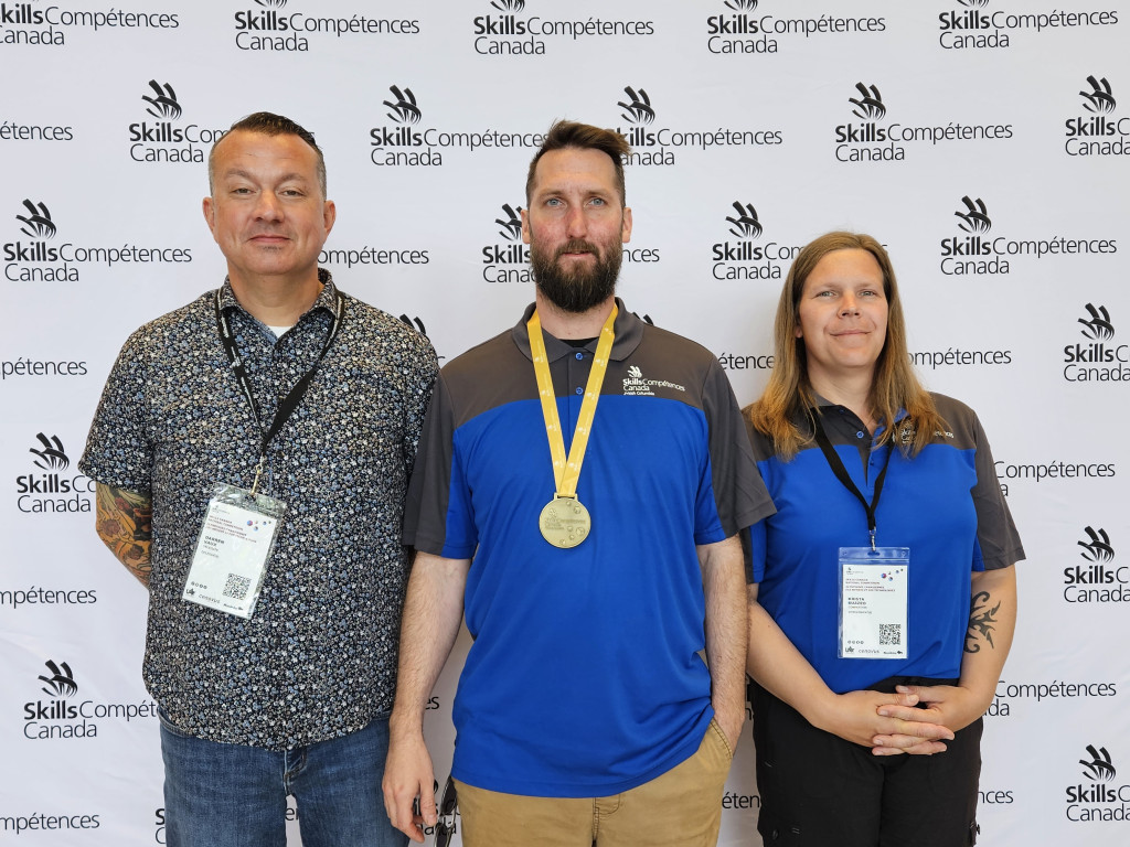 Three people pose against a Skills Canada backdrop. One older man in casual attire and two younger people wearing blue tops with the skills canada logo. The man in the middle has a gold medal around his neck.