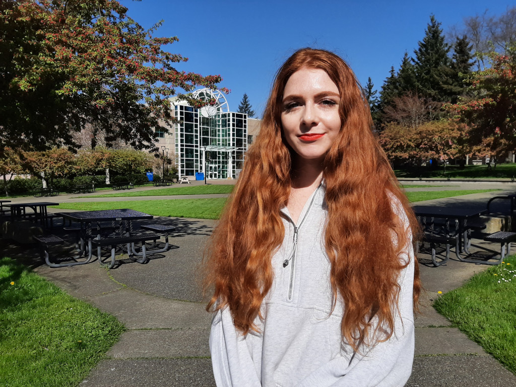 Portrait of a female student standing outside in the sunshine with Campus Centre on the Interurban campus in the backgroind
