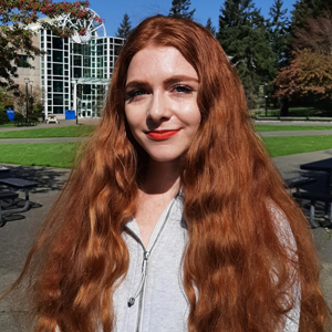 A young woman with long red hair standing in the sunshine outside of the student centre. 