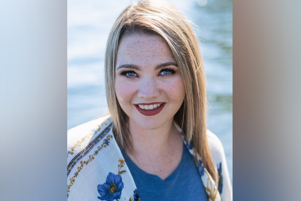 Portrait photograph taken outdoors featuring a young white woman. Backdrop looks like shimmering water but is out of focus. She is wear a blue v-neck top with a patterned cardigan.