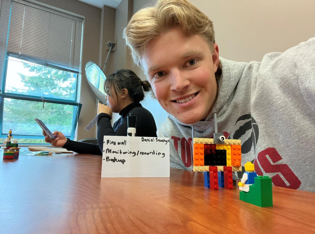 A male student with blonde haoir ooks at the camera showing off his lego model 