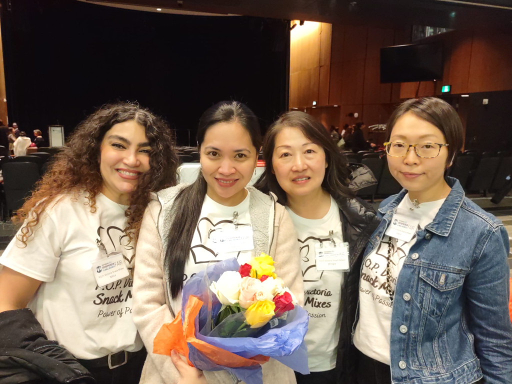 Four very happy looking women in a large indoor room pose for a group photograph. The space looks like a large auditorium. One woman is holding a colourful bouquet of flowers.