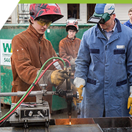 Two men in welding gear skillfully working on a metal object, creating sparks.