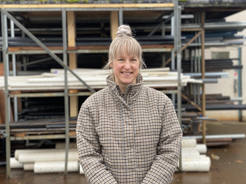 A woman with blonde hair and a check jacket stands in front of a rack of pipes.