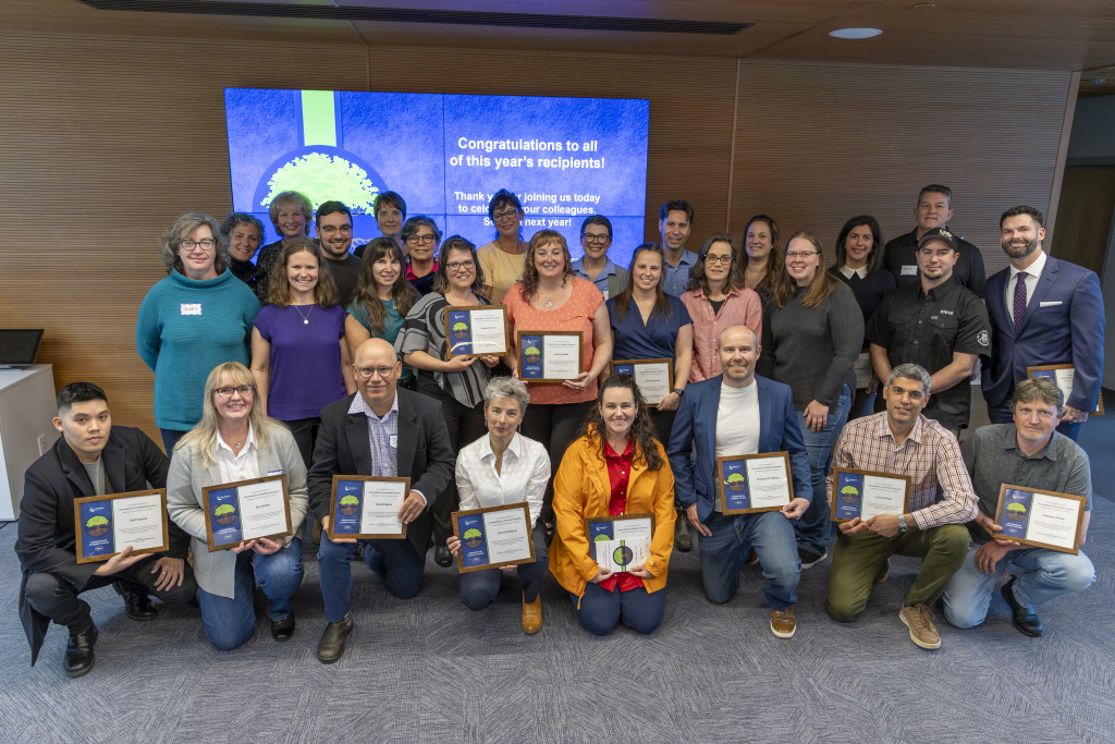 A mix of peoples pose indoors for a group photograph. The instructors are prouldy holding up their award certificates.