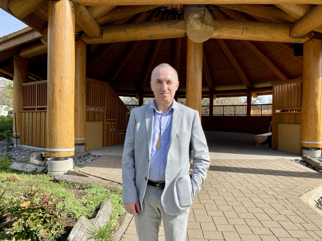 A man in a grey suit stands in front of a wooden building.