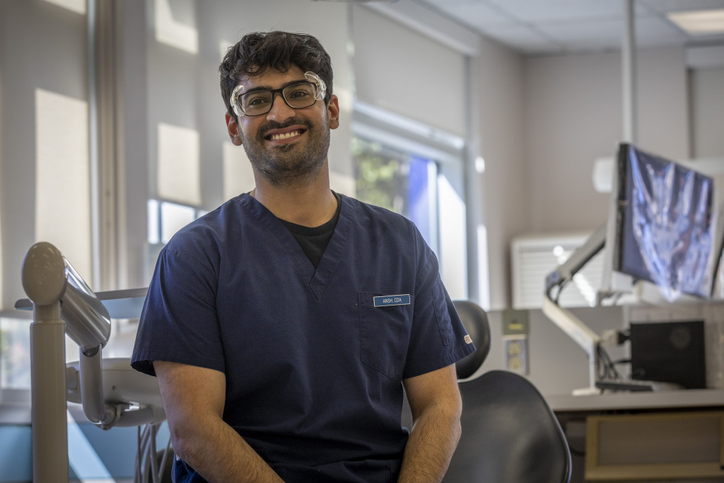 Ansh sitting in the dental clinic, wearing his scrubs and name tag, and safety glasses, smiling for the camera.