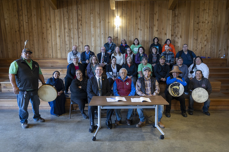 Members of Malahat Nation and Camosun College sit for a group of photo with a table and two signing document in the forefront