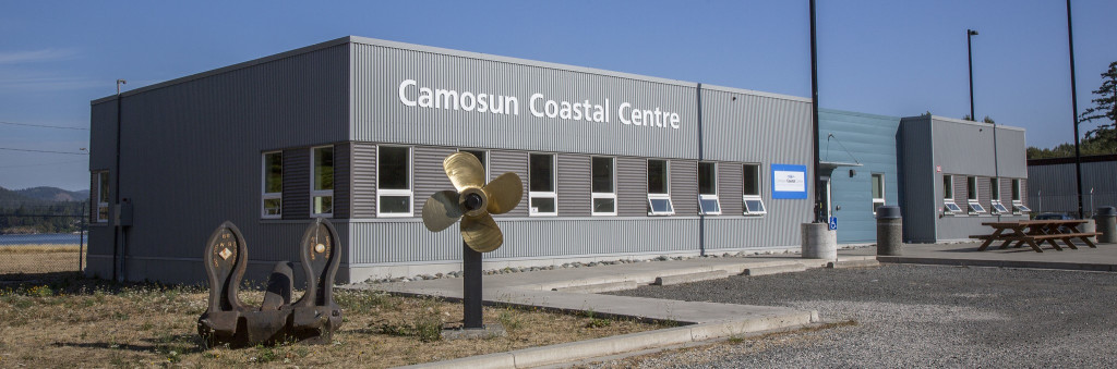 An industrial looking building with a the ocean and a shipyard in the background.  A large sign over the entrance says  sign that says "Camosun Coastal Centre".