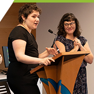 A student stands at a podium speaking to an audience. In the background a teacher looks on with a look a pride. 