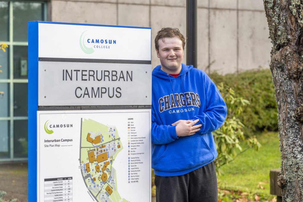 ETP student Jacob Rosser leans against an outdoor sign that says Interurban Campus. Jacob is wearing a royal blue hoodie saying Camosun Chargers.