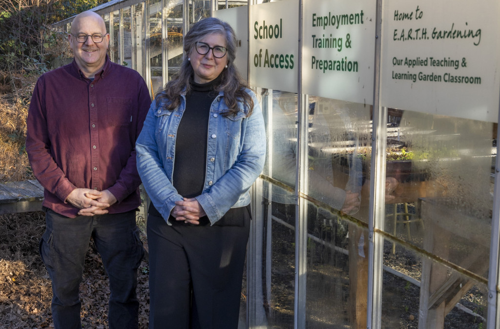 Instructors Mark Fournier and Laura Friesen stand next to each other with a glass greenhouse behind them. Signage on the side of the greenhouse says Employment Training and Preparation program with the college logo as well as saying Home to Earth Gardening.