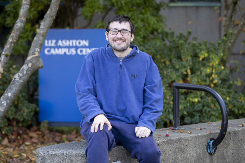 ETP grad Nic Ihmels sits on an outdoor concrete bench. He is wearing black glasses and a blue sweater. The background has a blue sign saying Liz Ashton Campus Centre.