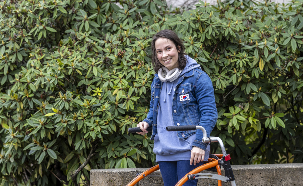 A young woman with dark hair and wearing a blue denim jacket poses for a photo. The background is a green shrub. The woman is holding onto an orange walker.