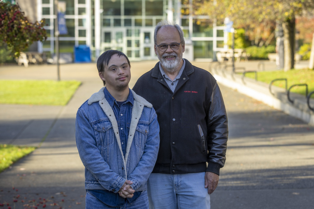 ETP graduate Evan Sturdy wearing denim jacket and jeans stands next to dad John Sturdy who is older with a beard.