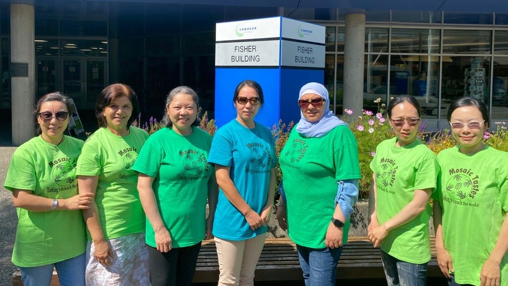 Seven women in green shirts stand in a row in front a blue sign that says "Fisher Building."