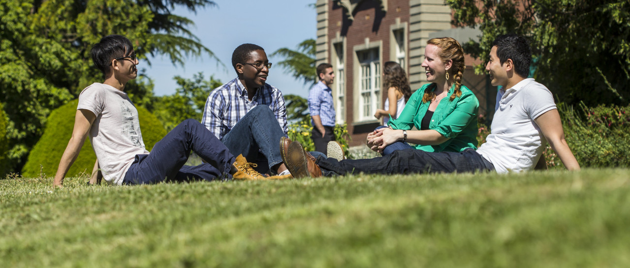 A group of 5 students sitting on the grass. They are talking and laughing.