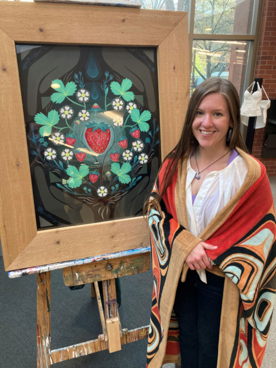 Student Alisha Parks wearing an Indigenous shawl stand next to her Heartberry painting