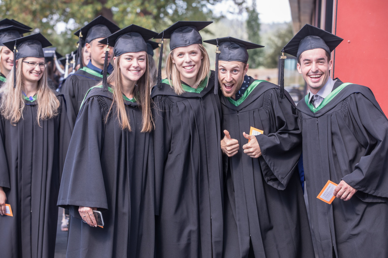 A group of young peple wearing black gowns with a green stripe around the collar and black caps stand next to each other to pose for an impromptu photograph.