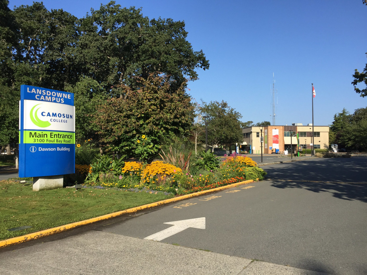 Image of Interurban campus sign for Camosun College on a nice sunny day with trees and a building in the background. Entry road with a white arrow in the front.