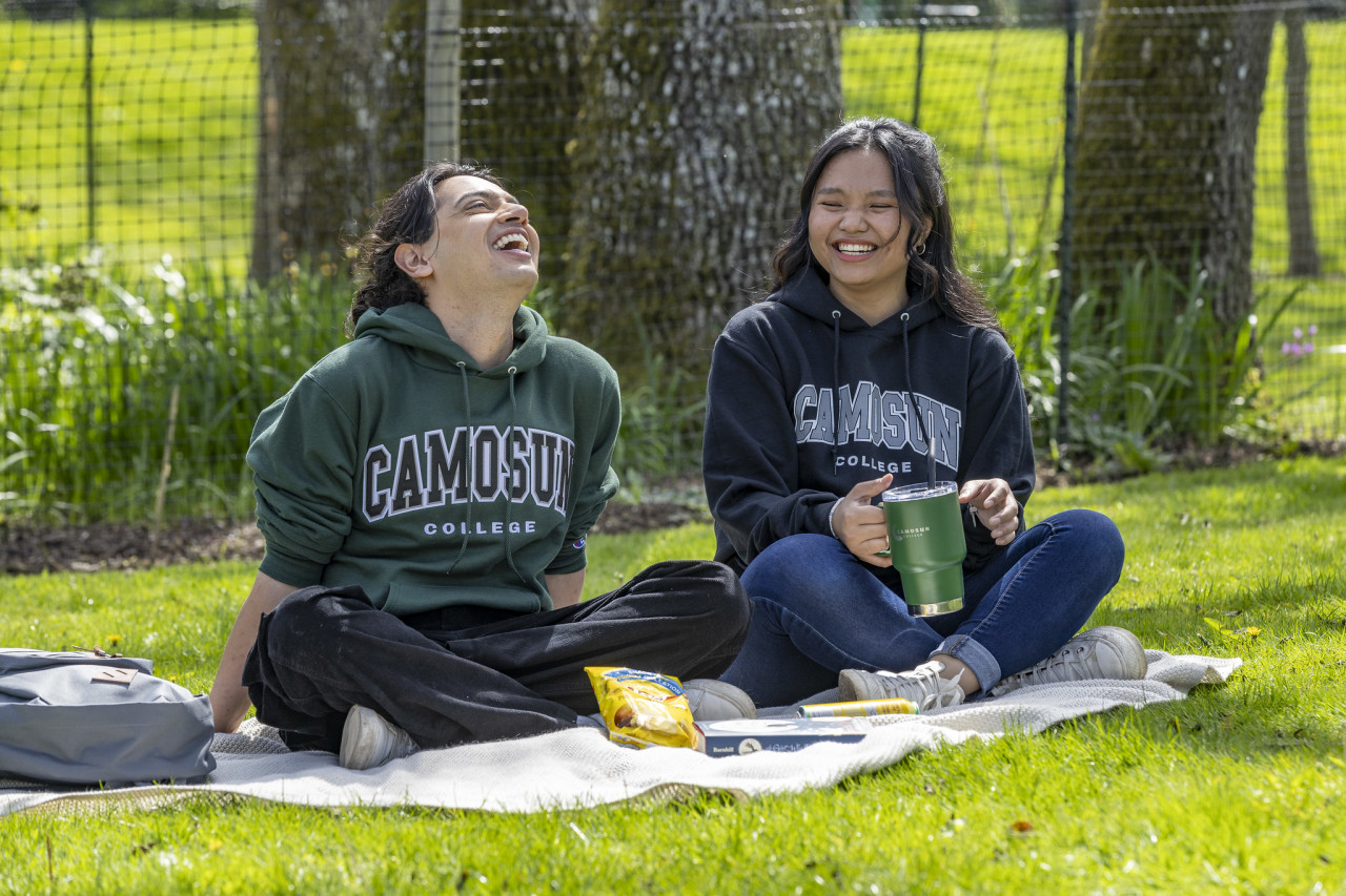 Two students sitting outdoors, having a picnic and wearing Camosun merch.