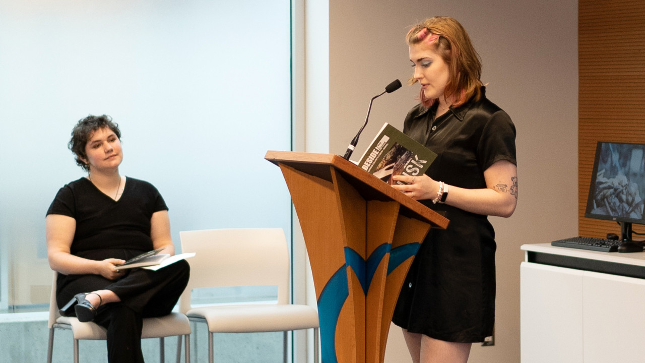 A women is at a podium reading from a book.  A second woman looks on from the side of the room.