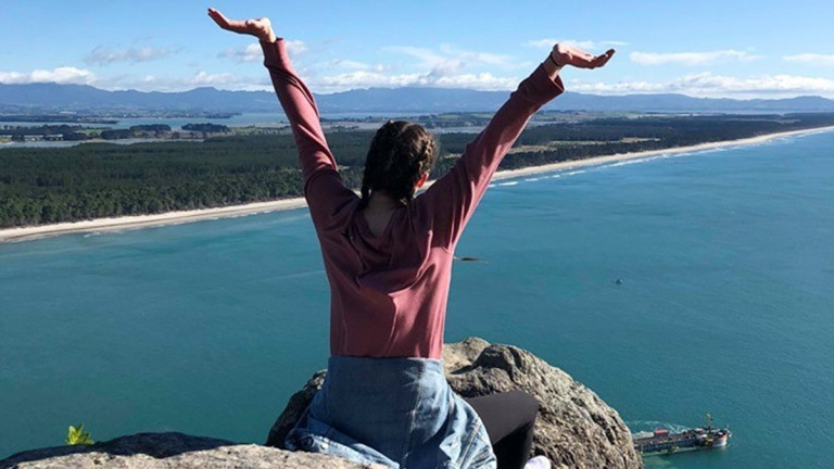 A woman raises her hands in joy as she sits on the edge of a cliff. 