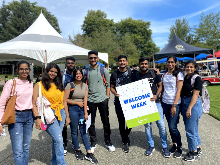 Nine students hold a large welcome sign. They are outside on campus. It's a sunny day and there is lots of activity happening in the background. 