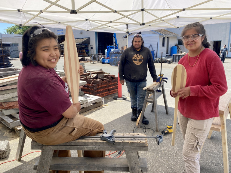 Three Indigenous students gather around woodworking equipment and tools.