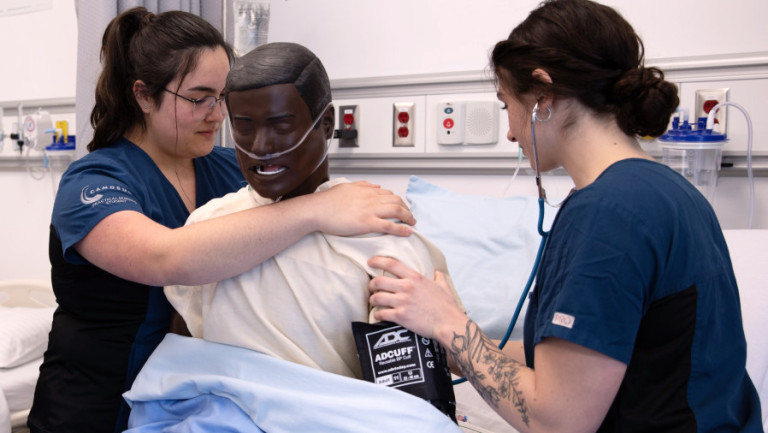 Two nursing students use a mannequin to practice helping patients sit up in bed.
