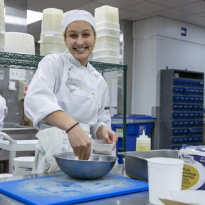A young women smiles and looks at the camera mixes ingredients while preparing food in a commercial kitchen.