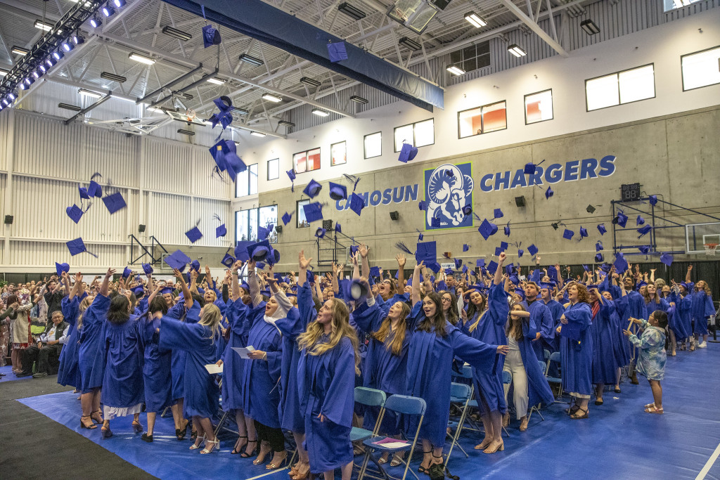 A large group of graduates wearing blue gowns are standing up as they toss blue cap above their heads