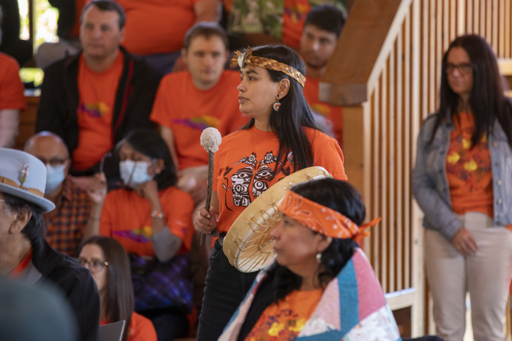 An Indigenous woman. Katie Manomie, in an orange shirt playing a drum in front of a gathering of people.