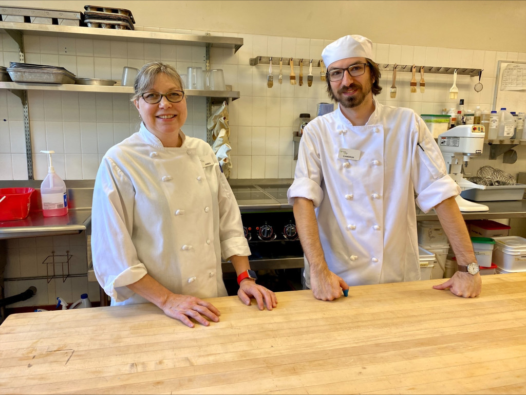 Two professional cook students wearing chef whites pose for a photograph in a commercial kitchen with their hands on a wooden bench.