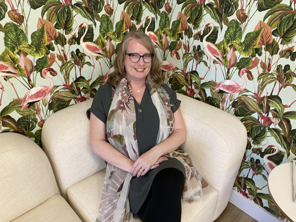 A woman in a green shirt sits on a couch with background wallpaper featuring flamingos and greenery